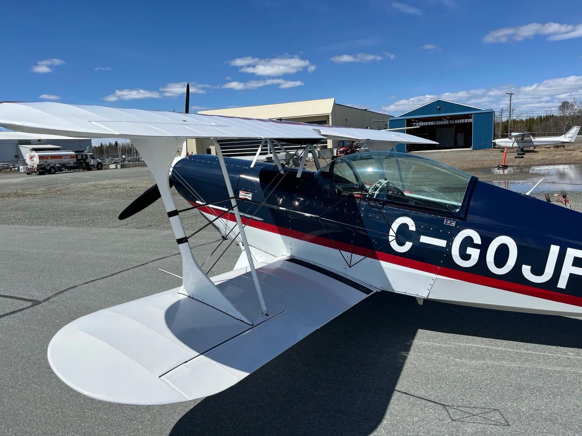A dark blue biplane with red and white stripes and white wings is parked on an airport tarmac under a blue sky.
