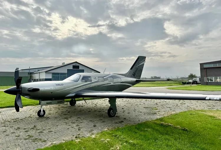 A Piper M600 HALO aircraft parked on the grass in front of a hangar on a cloudy day.