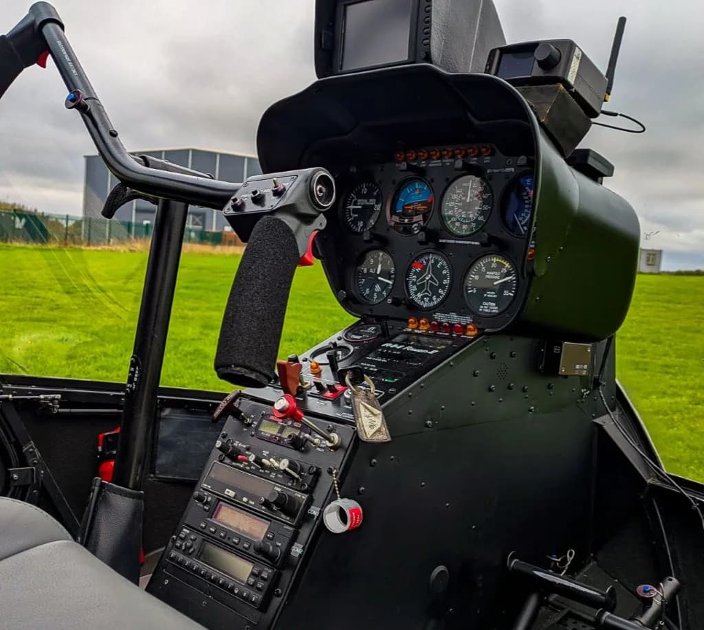 The instrument panel and cyclic control of a helicopter, with a green field visible through the windshield.