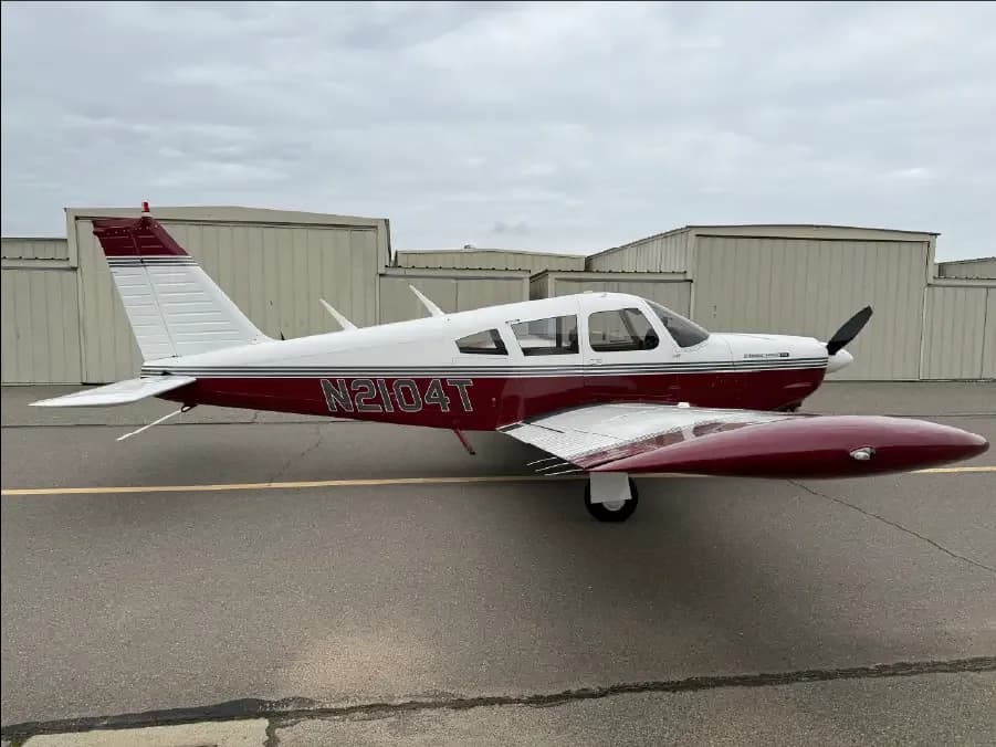A white and maroon single-engine propeller aircraft, N2104T, is parked on an asphalt ramp with hangars in the background under a cloudy sky.