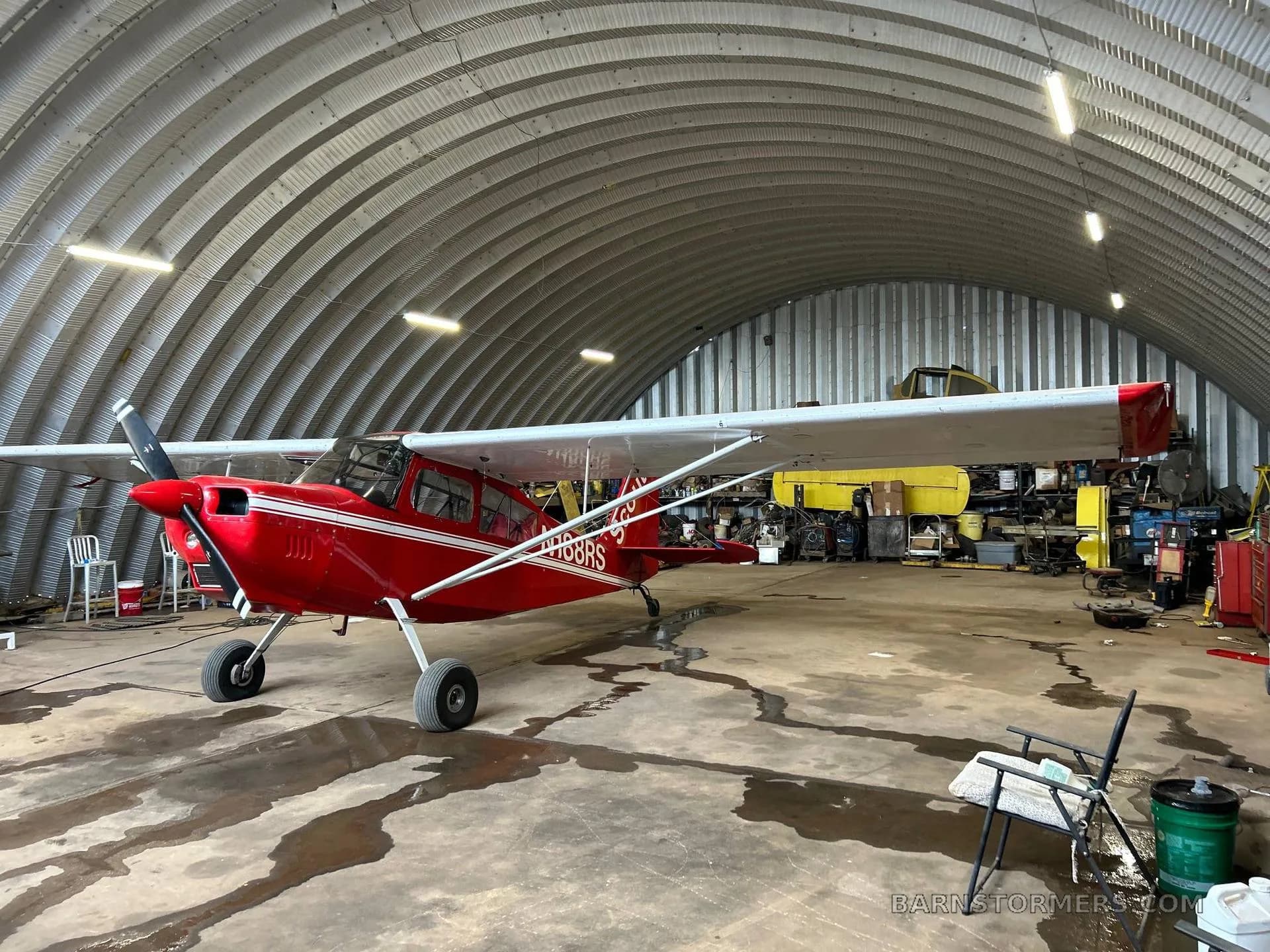 A red Scout N188RS aircraft with white stripes is parked inside a metal hangar, viewed from the front-left.