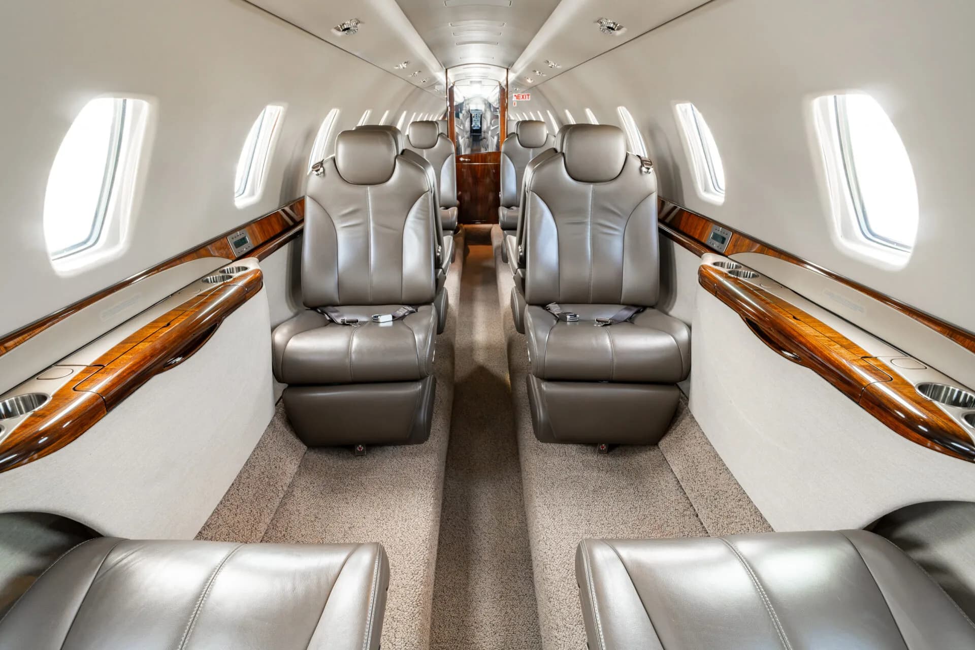 A view down the aisle of a business jet cabin showing rows of tan leather seats and large windows.