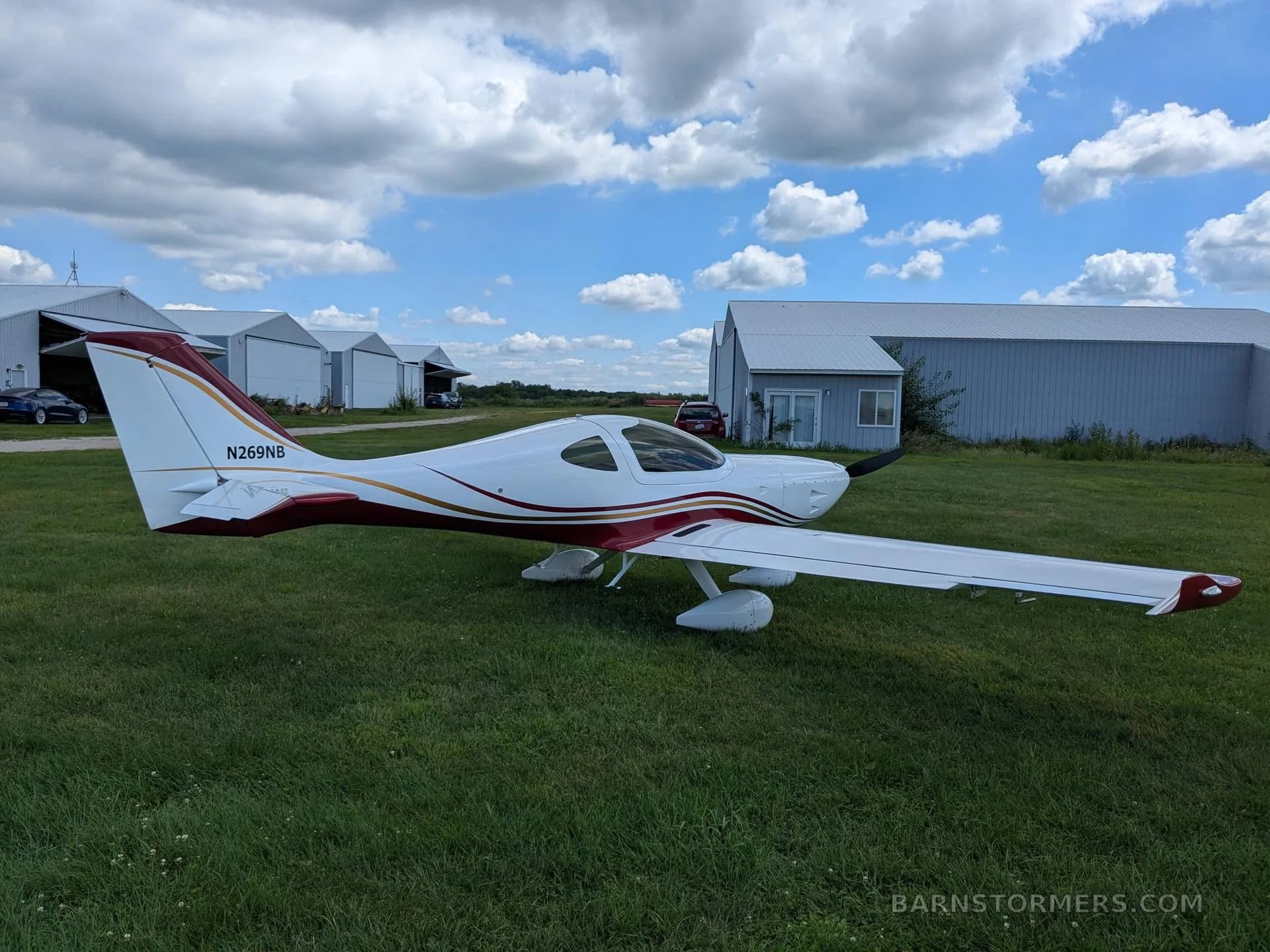 A white and red aircraft with tail number N269NB is parked on a grassy field with hangars in the background.
