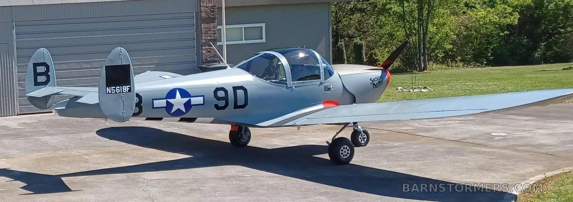 A grey single-engine aircraft with a red nose and "Sassy Baby" text is parked on a concrete driveway, viewed from the rear-right.