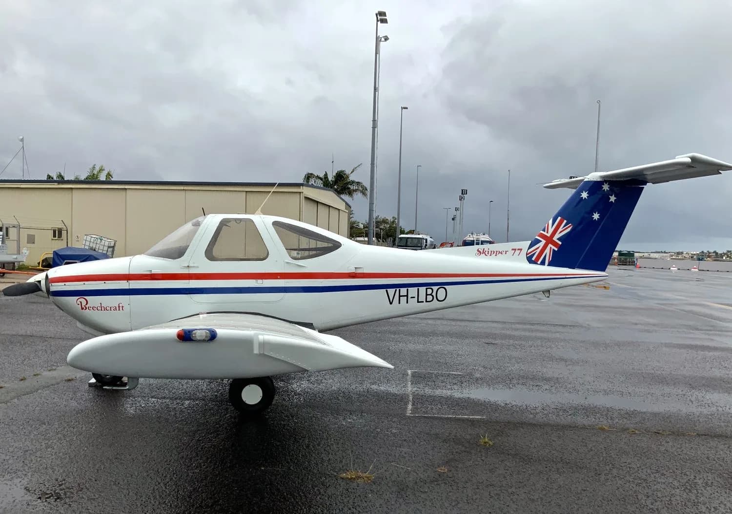 A white Beechcraft Skipper 77 with red and blue stripes and Australian flag tail, registration VH-LBO, parked on a wet tarmac under a cloudy sky.