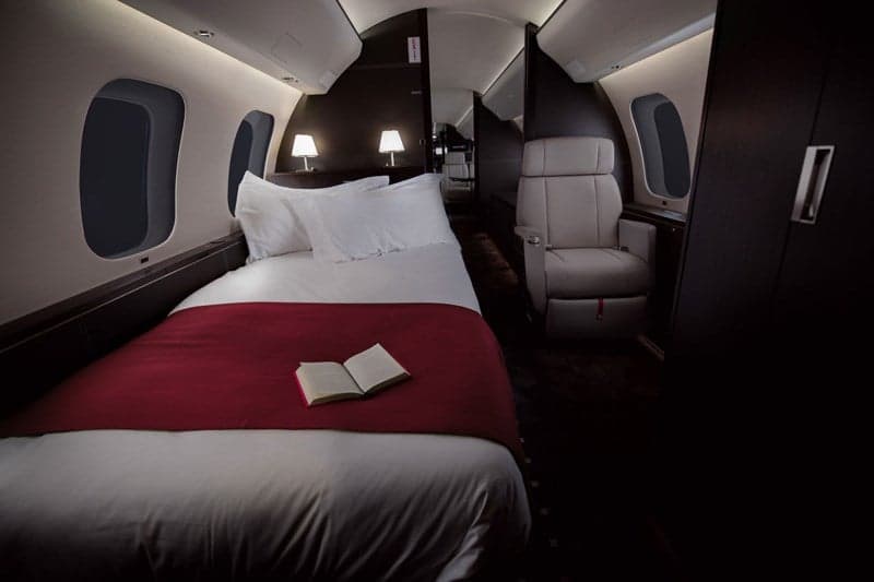 A private jet cabin interior featuring a comfortable bed with white sheets and a red blanket, alongside a single passenger seat.