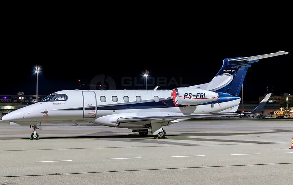 A white and blue Embraer Phenom 300E business jet with registration PS-FBL is parked on an airport tarmac at night.