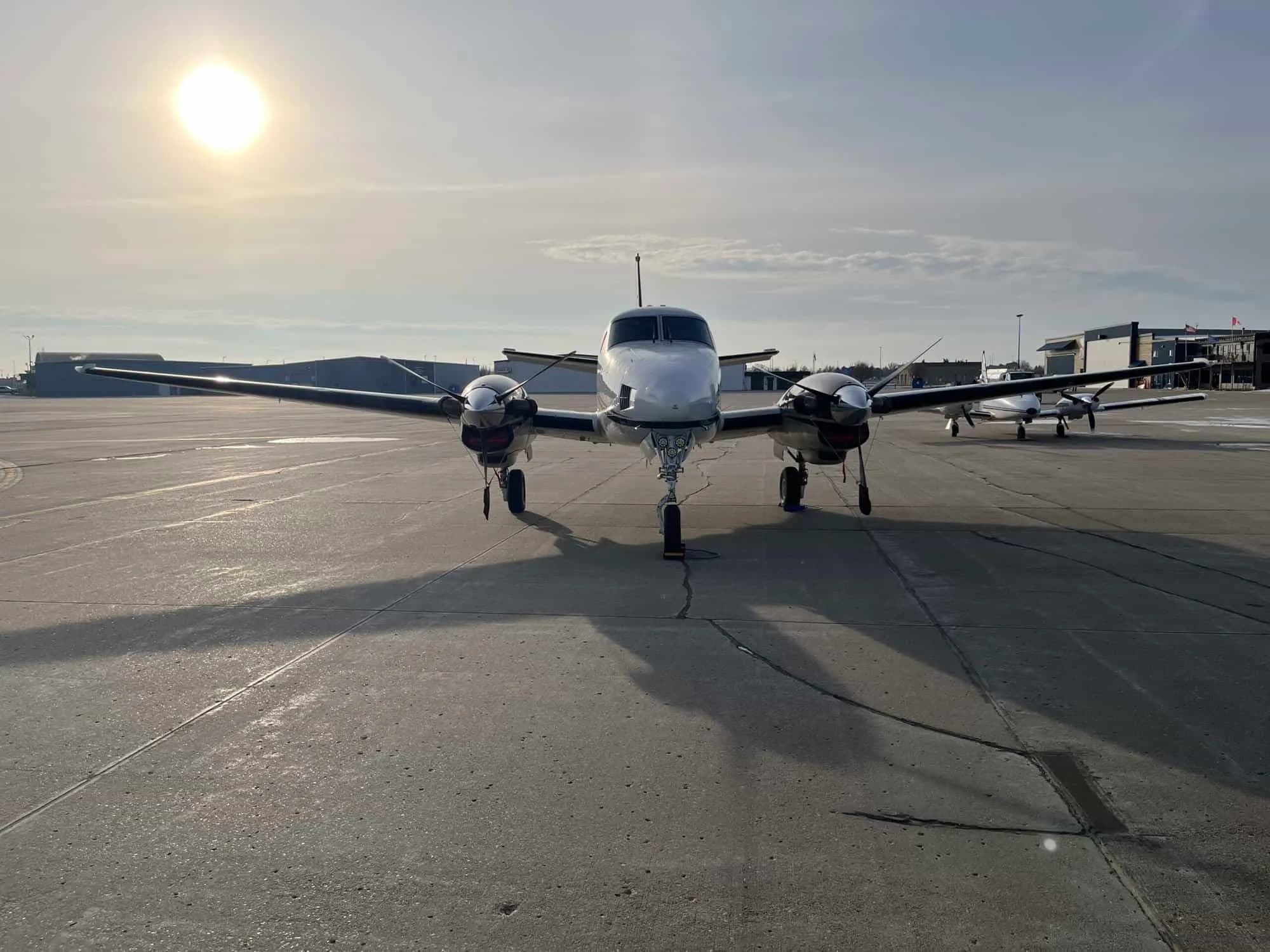 A head-on view of a white twin-engine turboprop aircraft with black propellers, parked on an airfield under a sunny sky.