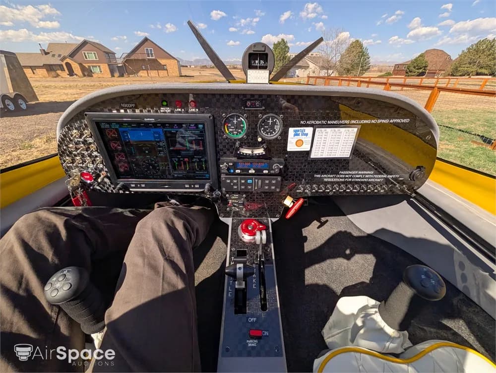 The cockpit of a yellow aircraft shows a modern glass panel display, traditional gauges, and flight controls, with a view of houses and trees outside.