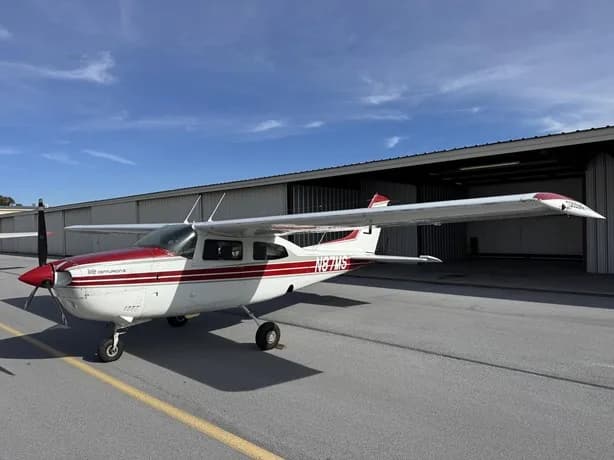 A white and red Cessna 210 Centurion aircraft, N87MS, is parked on an asphalt tarmac next to an open hangar under a blue sky.