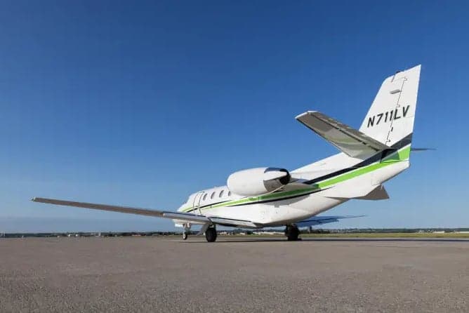 A white business jet with green and black stripes on the tail and fuselage, seen from the rear-right on an airfield under a clear blue sky.