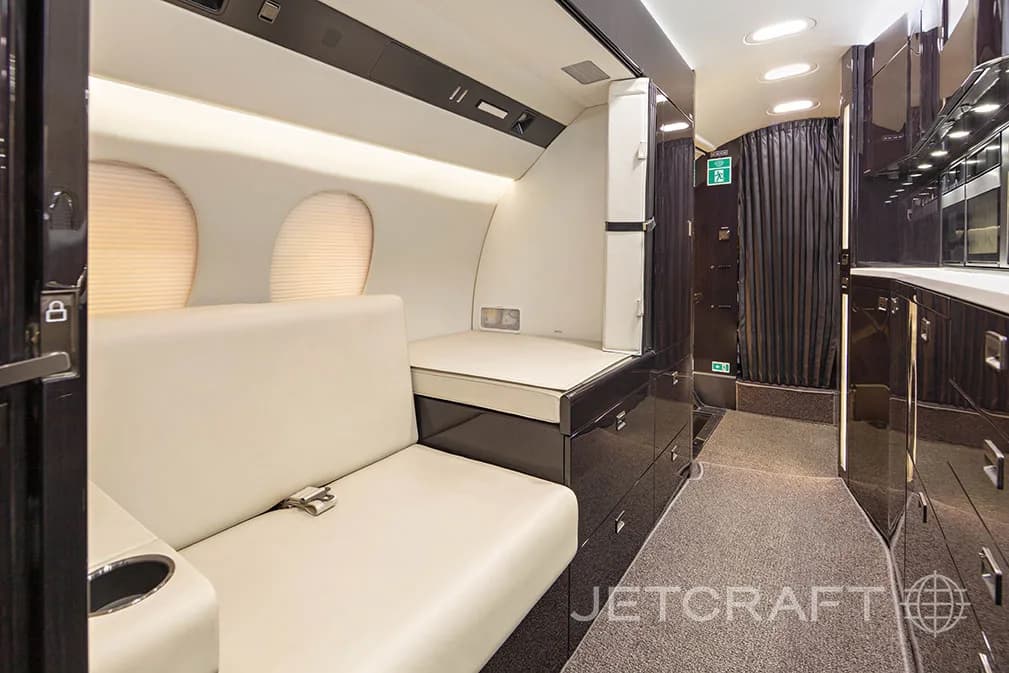 An aircraft cabin section with a cream-colored couch and a flat sleeping surface, next to dark wood cabinetry.