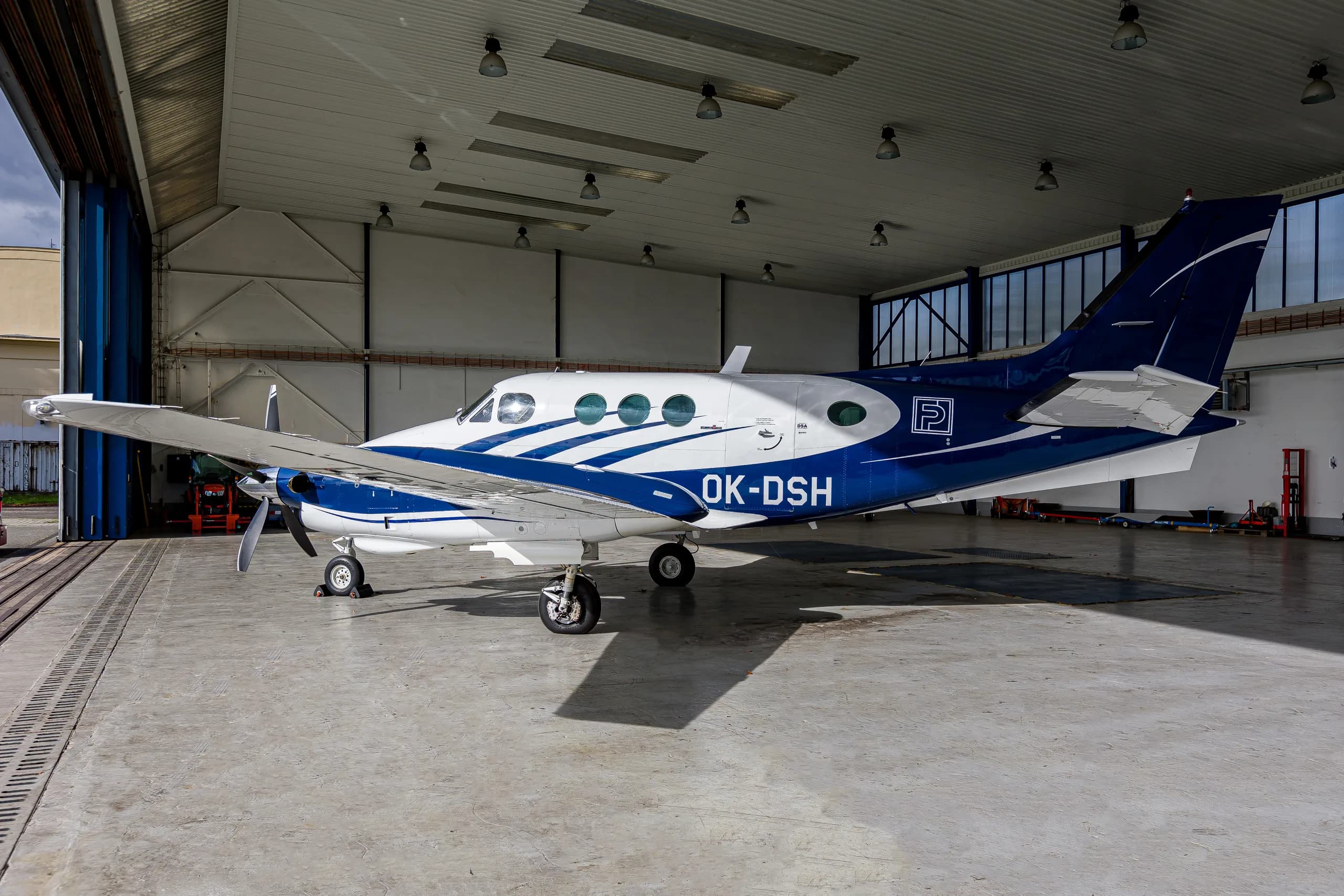 A white and blue twin-engine turboprop aircraft, registration OK-DSH, parked inside a hangar with its door open.