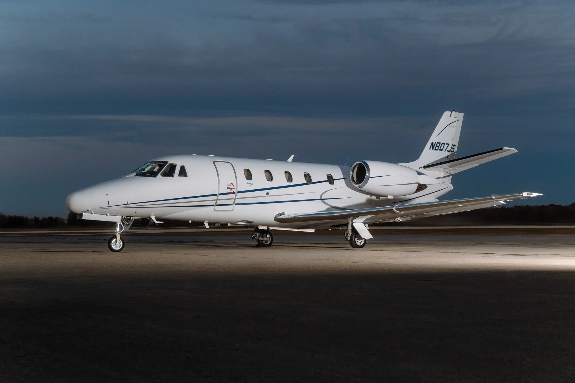 A white Cessna Citation X business jet with blue and grey stripes is parked on an airfield at dusk.