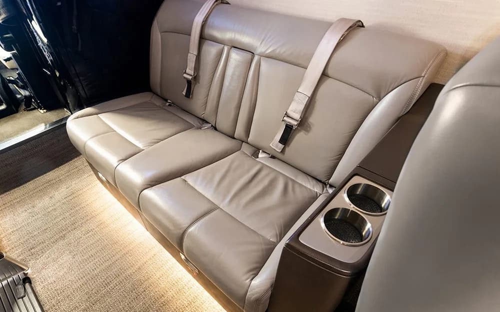A close-up of a light brown leather bench seat with seatbelts and cup holders in a private jet cabin.