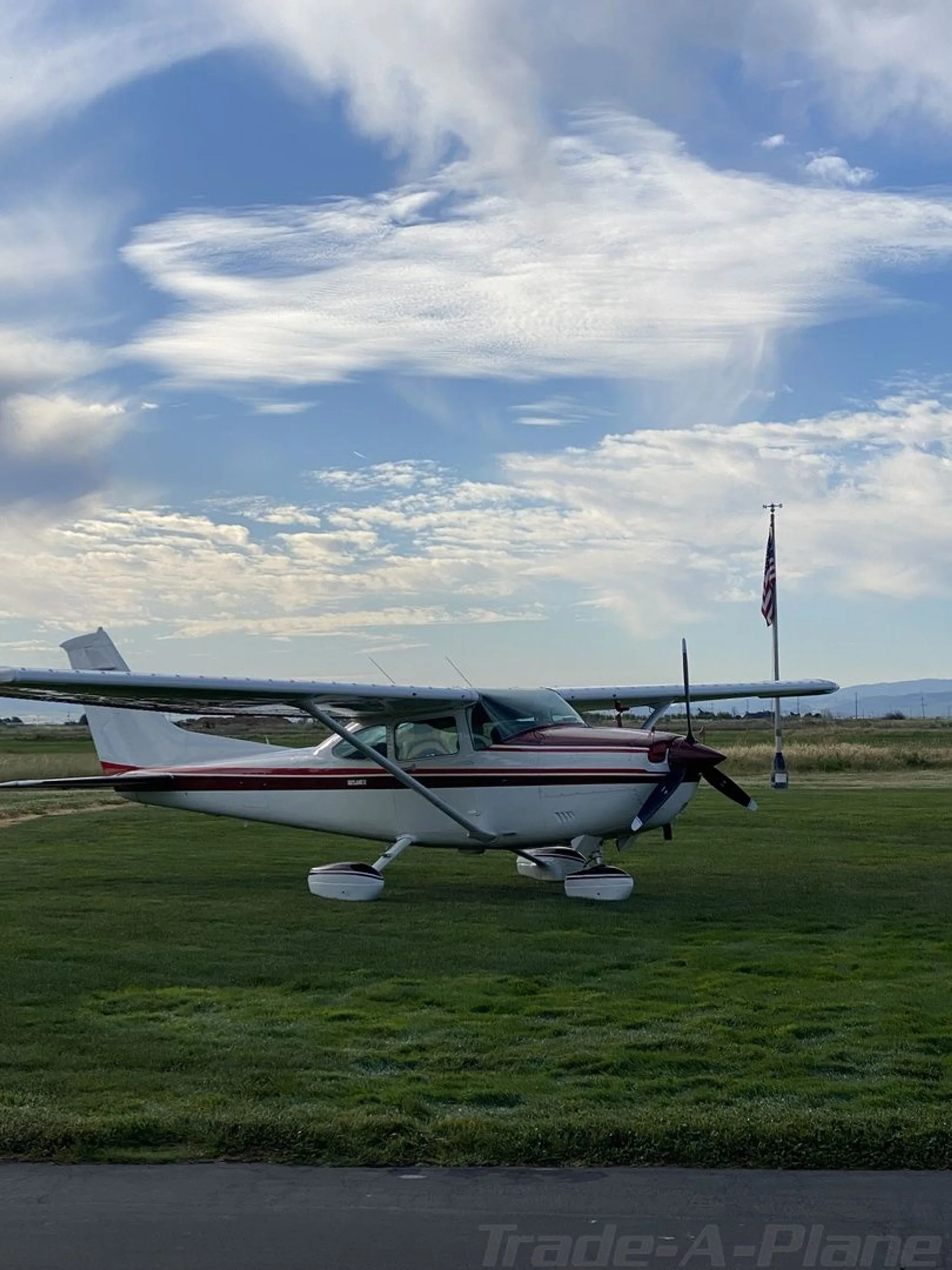 A white Cessna 182 with a red stripe sits on a grassy field under a cloudy sky, with an American flag visible in the background.