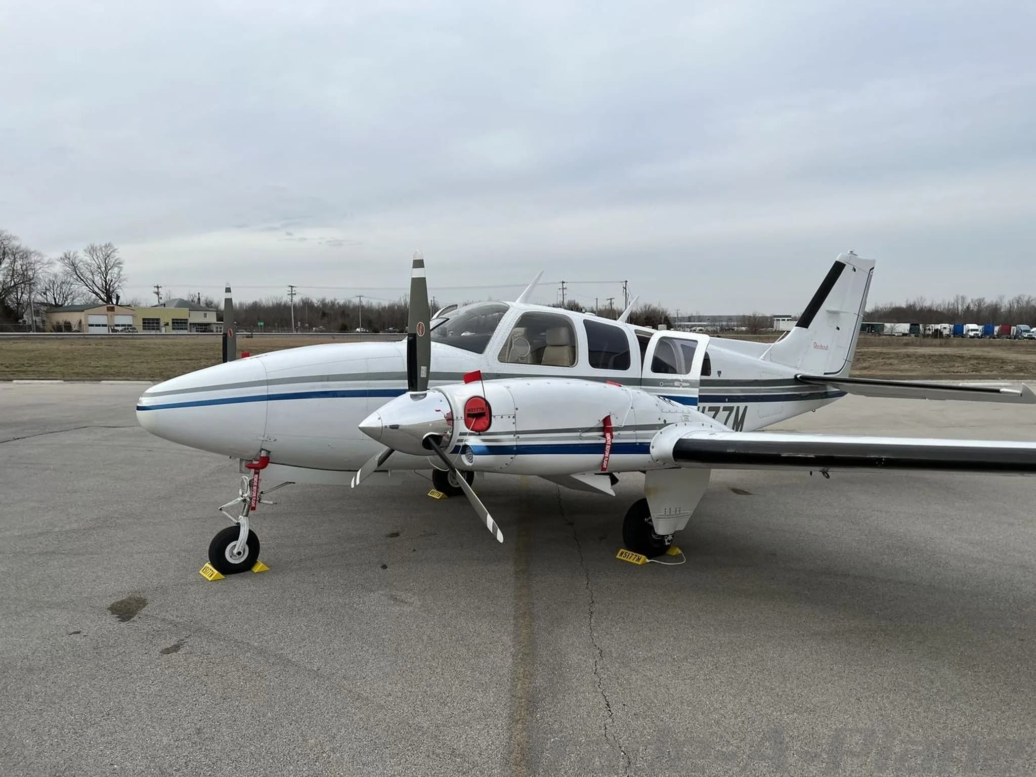 A white and blue twin-engine Beechcraft Baron N5177M parked on an asphalt ramp under an overcast sky.