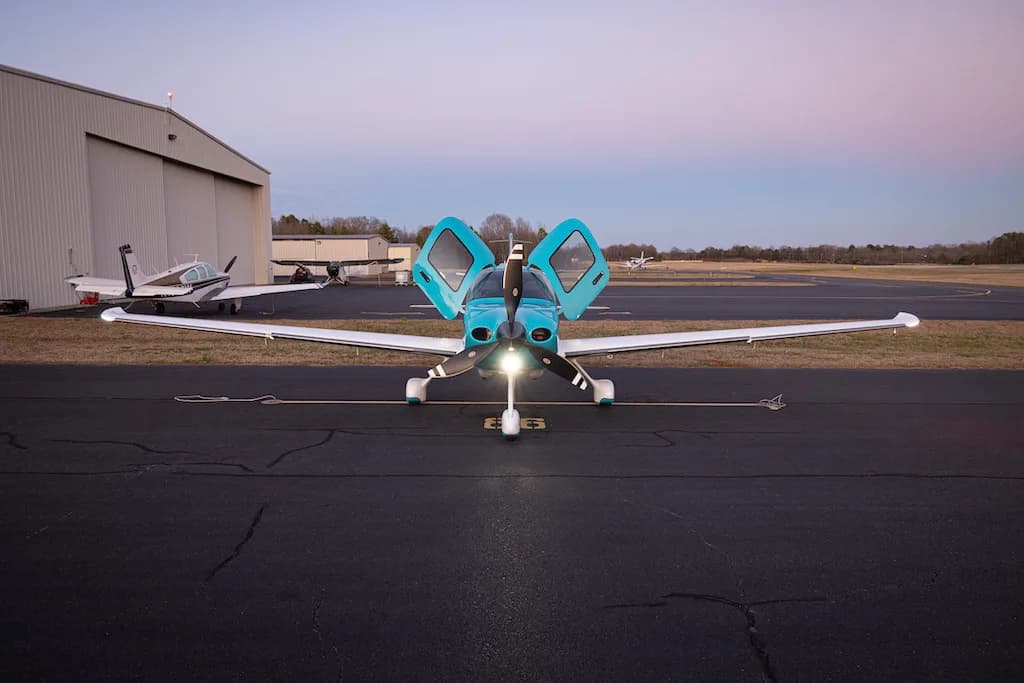 A front view of a turquoise and white Cirrus SR22 aircraft with its doors open, parked on an airfield at dusk.