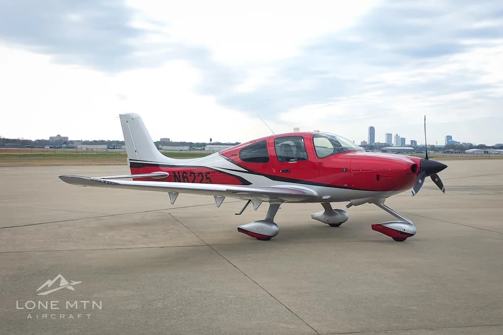 A red and silver Cirrus SR22 aircraft, N6225, parked on an airport apron with a distant city skyline.