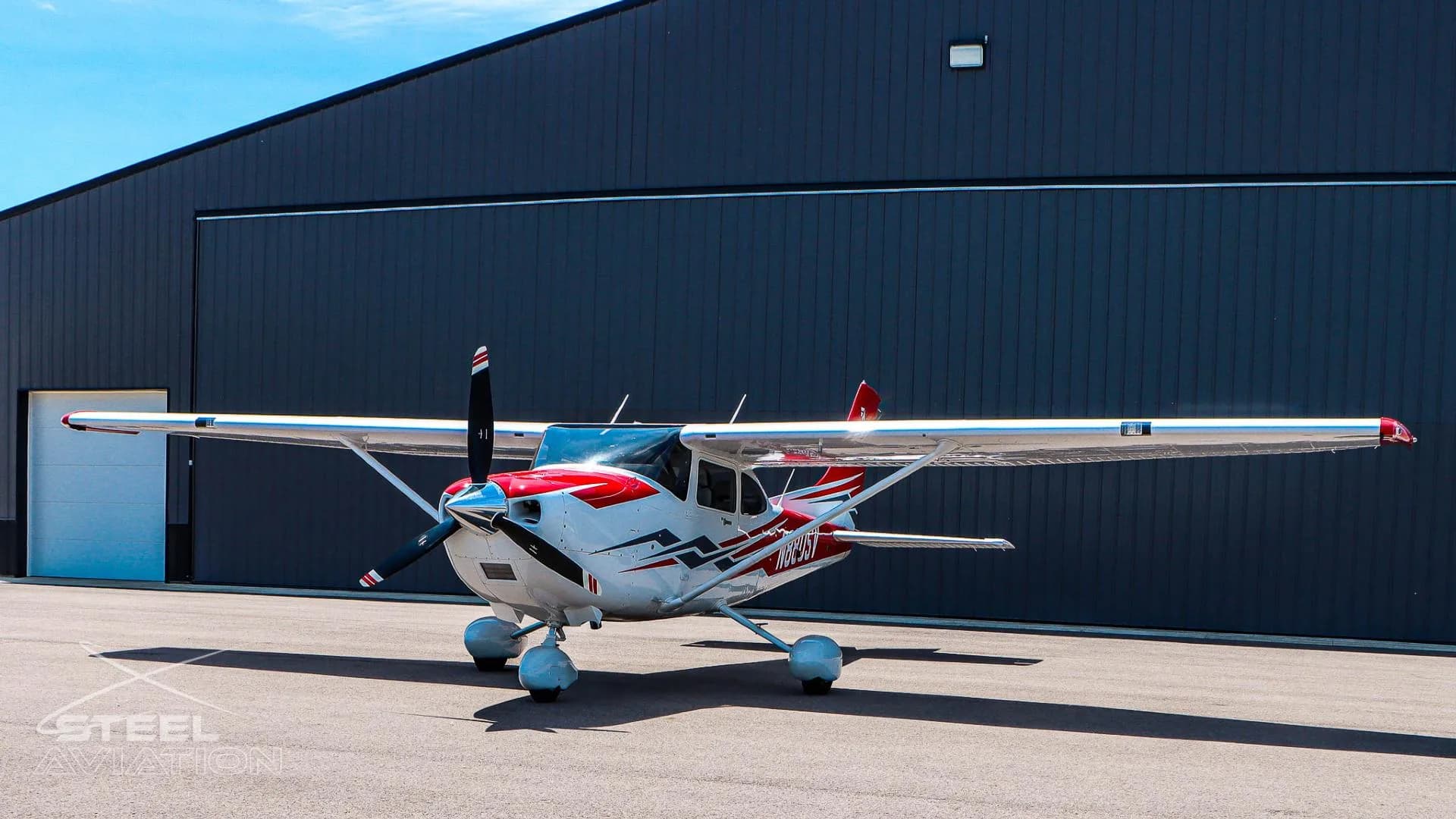 A three-quarter front view of a white Cessna T182T Skylane TC with red and grey stripes, parked on an asphalt tarmac next to a dark hangar.