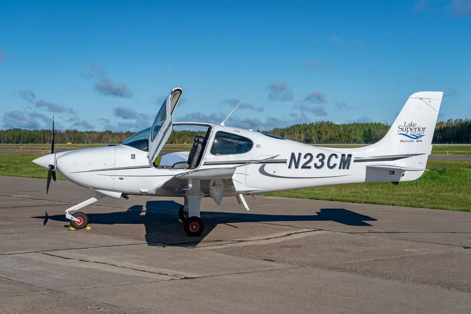A white Cirrus SR20 aircraft with registration N23CM and "Lake Superior College" logo on the tail, parked on an airfield with doors open.