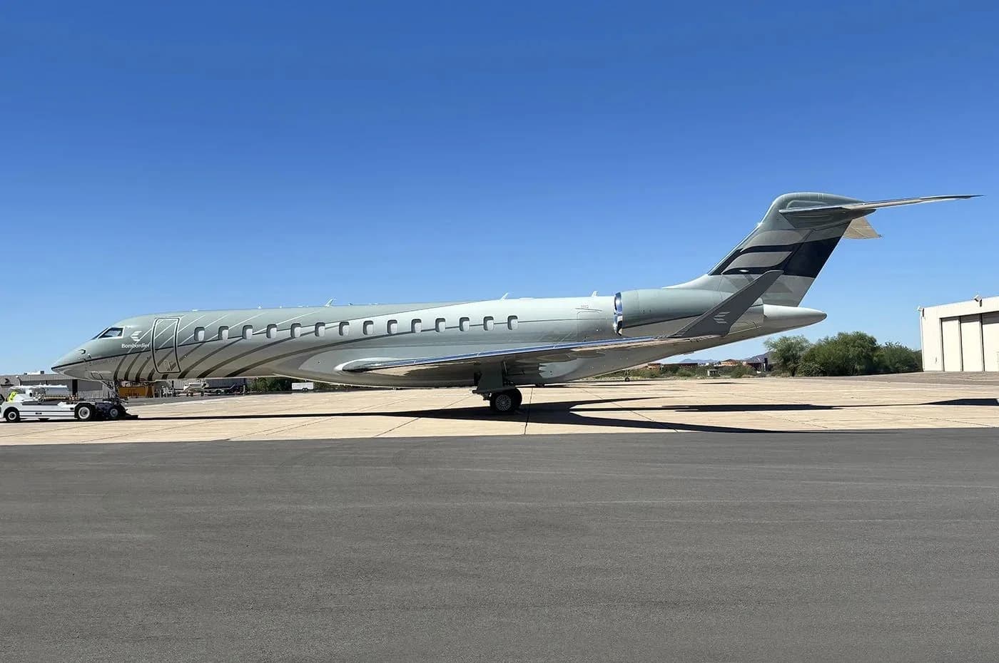 A grey private jet with a distinctive wavy paint scheme on the fuselage and a dark grey tail, parked on an asphalt tarmac under a clear blue sky.