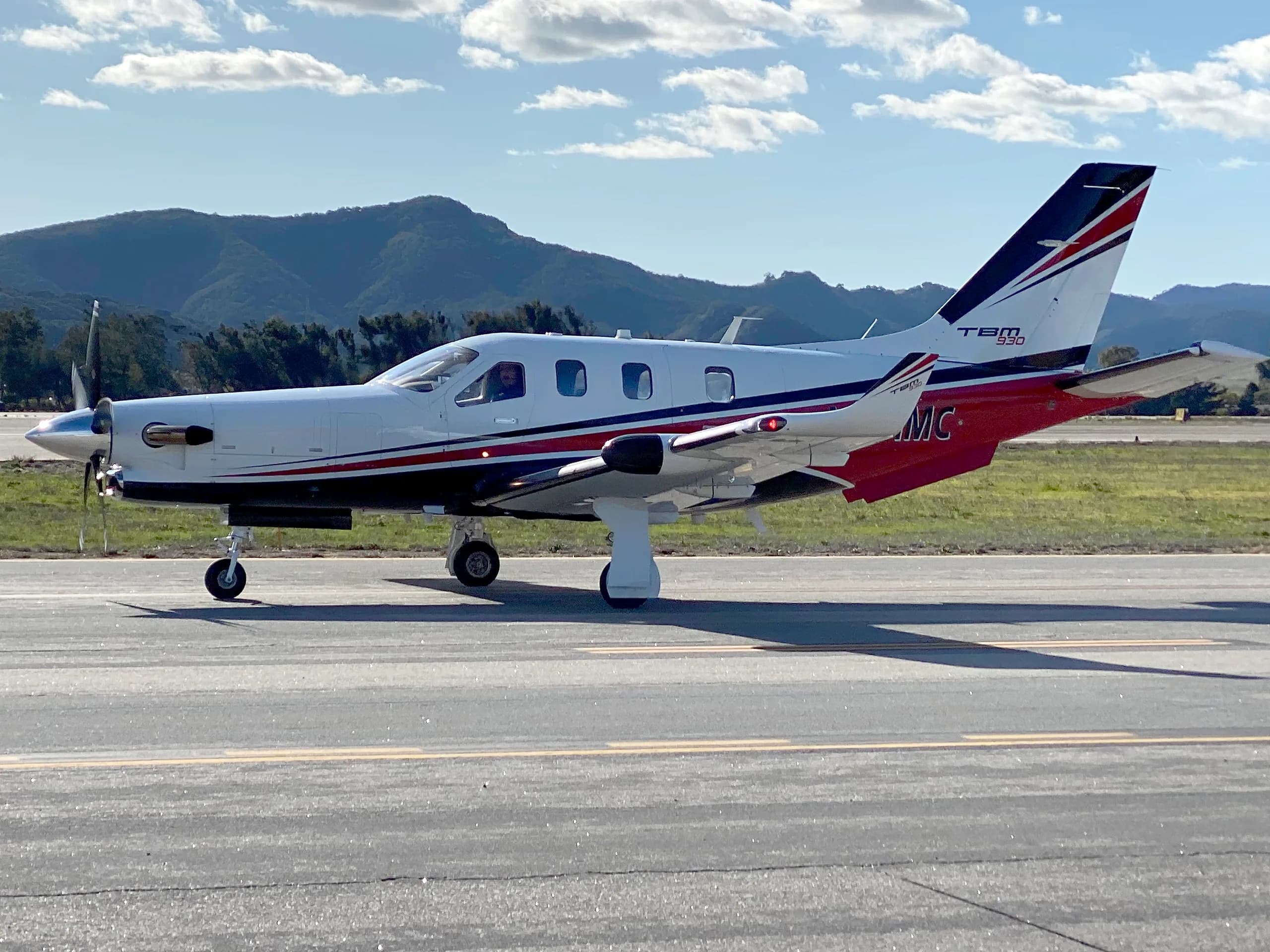 A white Daher TBM 930 aircraft with red and black stripes is seen from the side on an airfield, with mountains in the background.