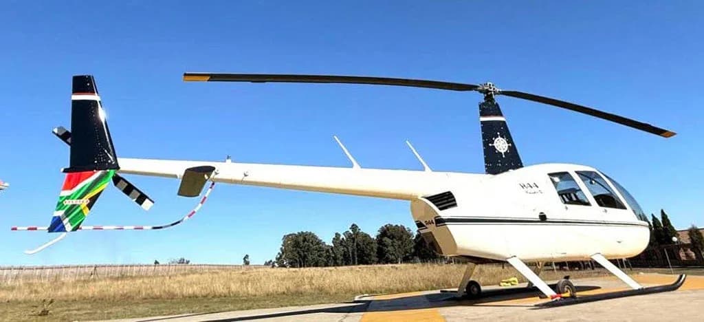 A white Robinson R44 Raven II helicopter with a South African flag on its tail boom is parked on a tarmac under a clear blue sky.
