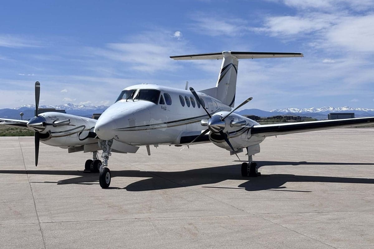 A white Beechcraft King Air N31WC parked on an airfield with snow-capped mountains in the distance.