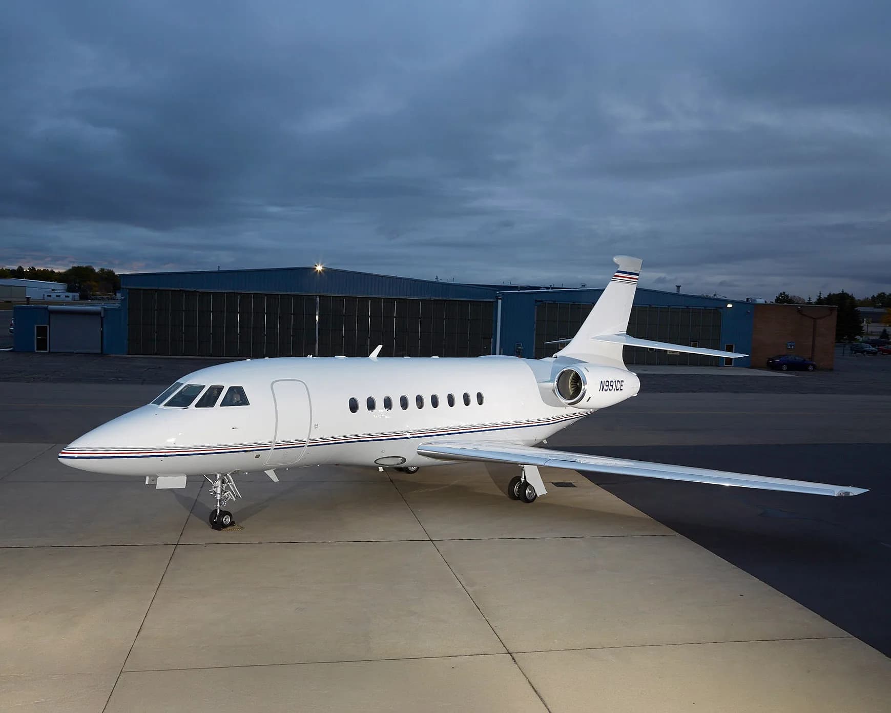 A white Dassault Falcon business jet with blue and red stripes on the tail and fuselage, parked on an tarmac with hangars in the background under a cloudy sky.