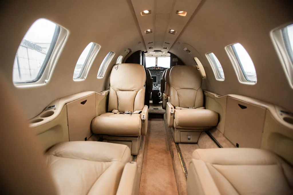 View of a private jet's beige leather cabin interior looking towards the cockpit, with two rows of seats visible.