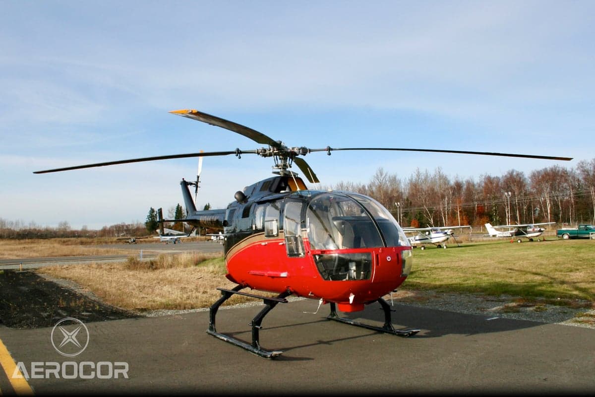 A red and black MBB BO-105 helicopter parked on an airfield with other aircraft and trees in the background.