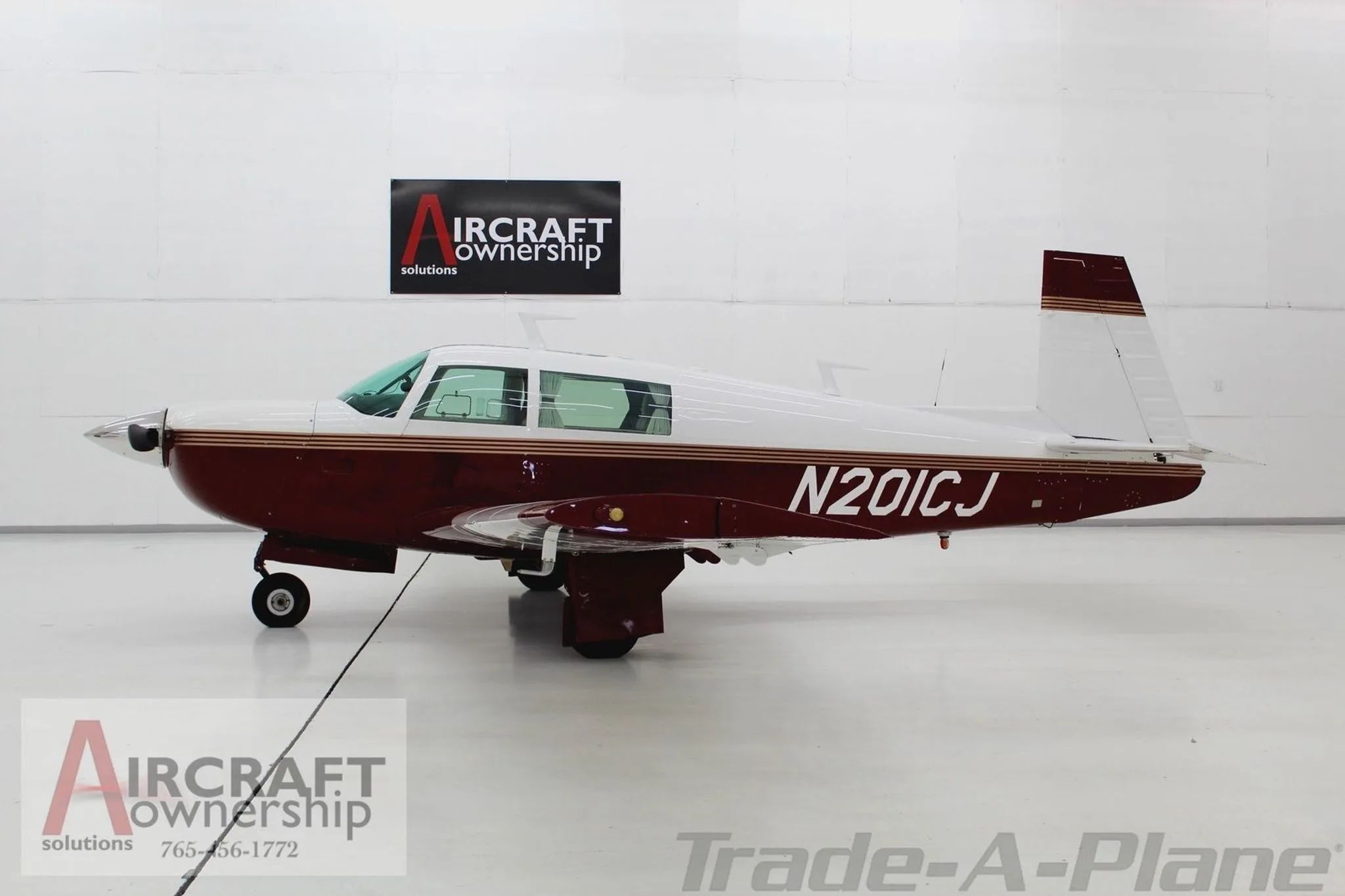 A side view of a Mooney M20J aircraft, N201CJ, in a hangar, featuring a white and maroon paint scheme with gold stripes.