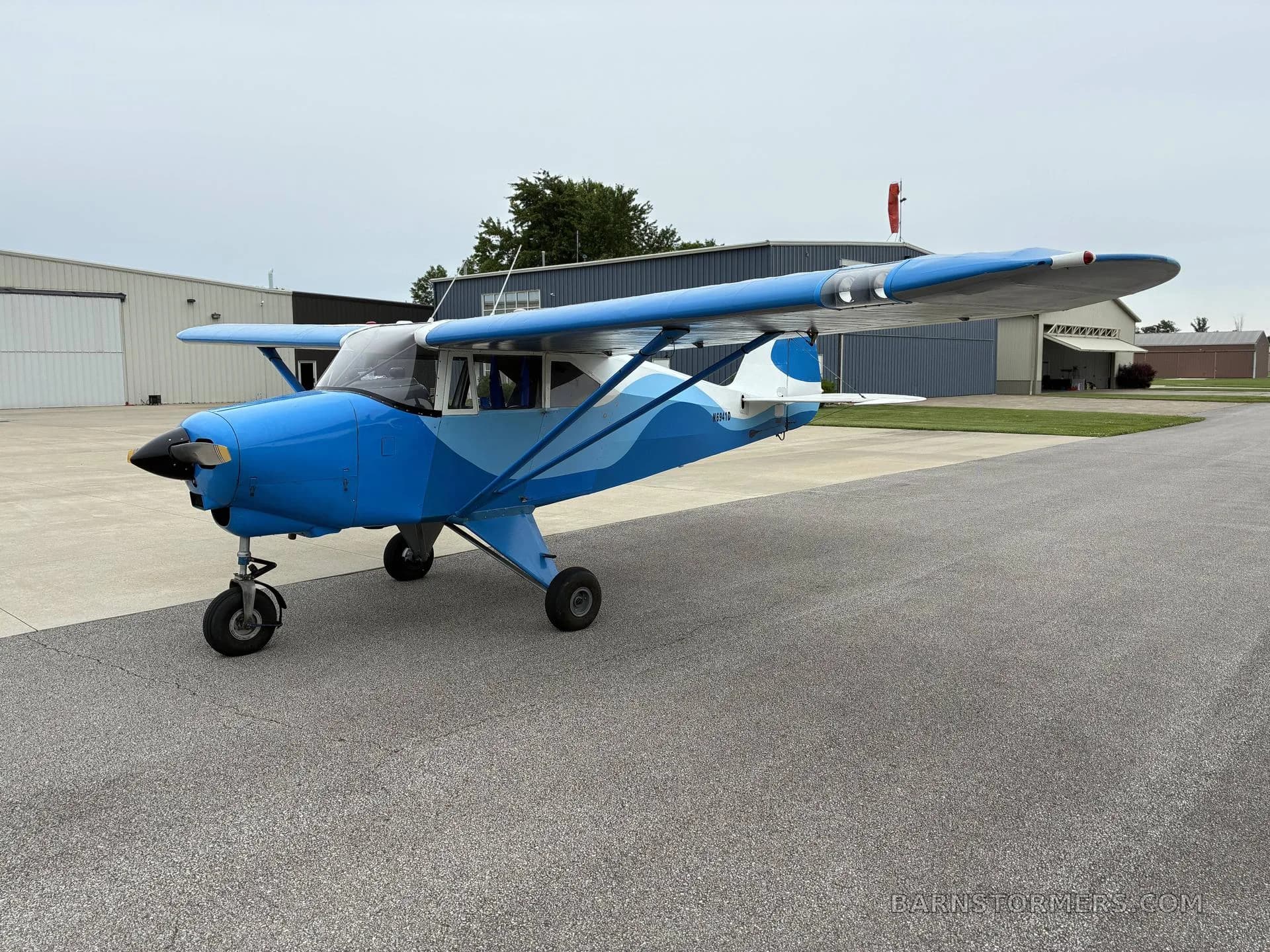 A blue and light blue Piper Cub-style aircraft with a wavy paint scheme is parked on an asphalt ramp in front of hangars under a cloudy sky.