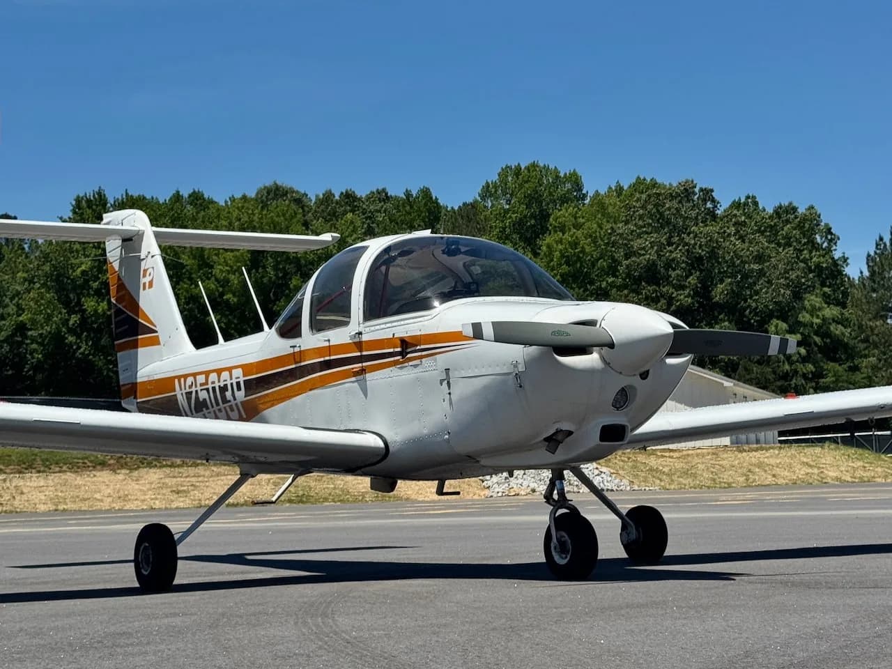 A white Piper PA-38 Tomahawk aircraft with orange and black stripes, seen from the front-right on an airfield.