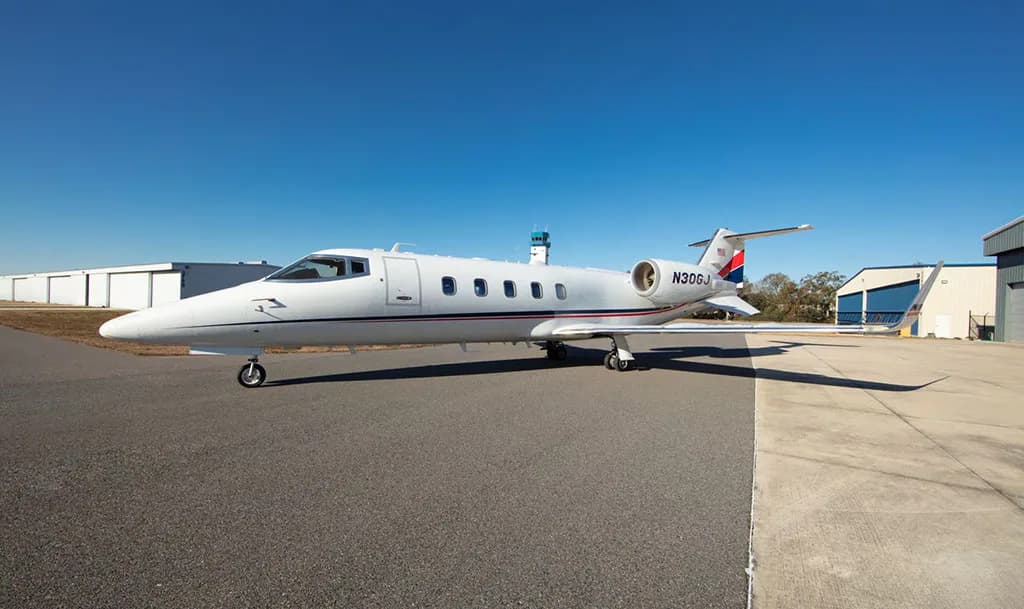 A white Learjet with red and blue tail stripes, registration N30GJ, parked on an airfield with a small control tower in the background.