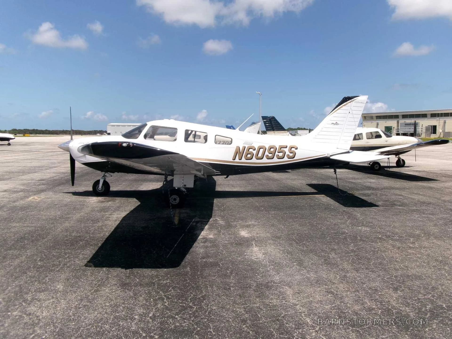A white and black Piper PA-28 Cherokee with gold trim, registration N6095S, parked on an asphalt ramp under a partly cloudy sky.