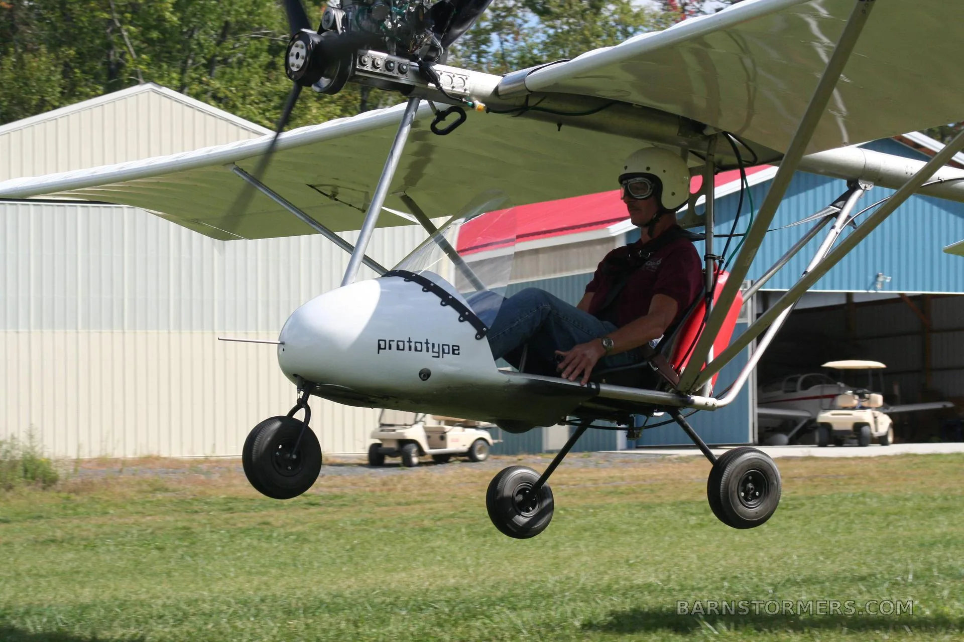A white ultralight aircraft with 'prototype' written on its nose, carrying a pilot, is shown just above a grassy field with hangars in the background.