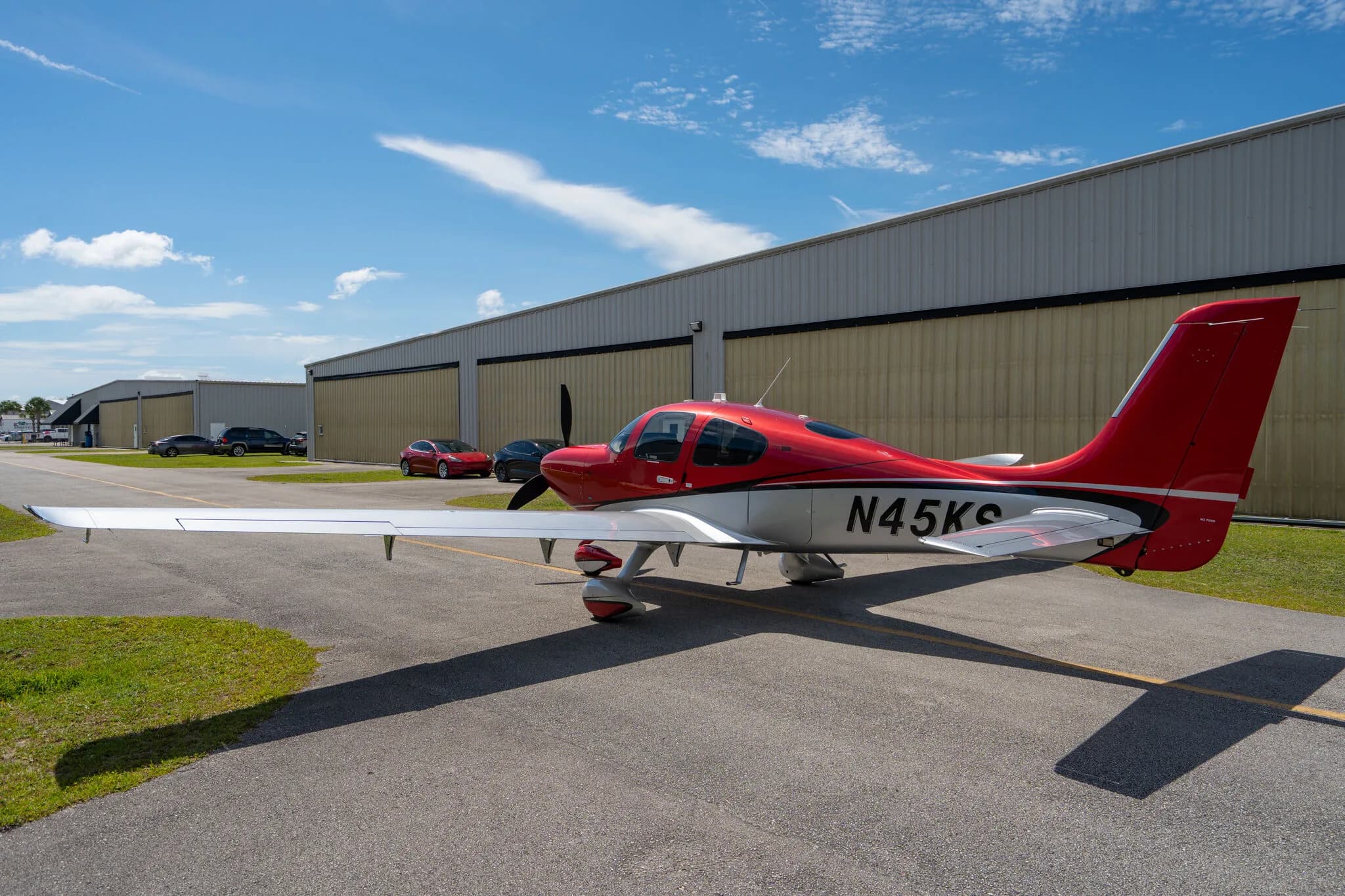 A red and silver Cirrus SR22 GTS (N45KS) parked on an airfield with hangars in the background.