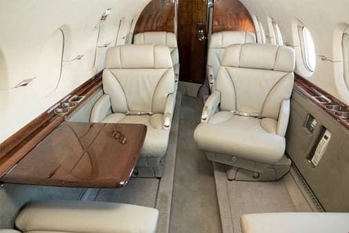 An aircraft cabin interior with light leather seats, a polished wooden table, and wood paneling along the walls.