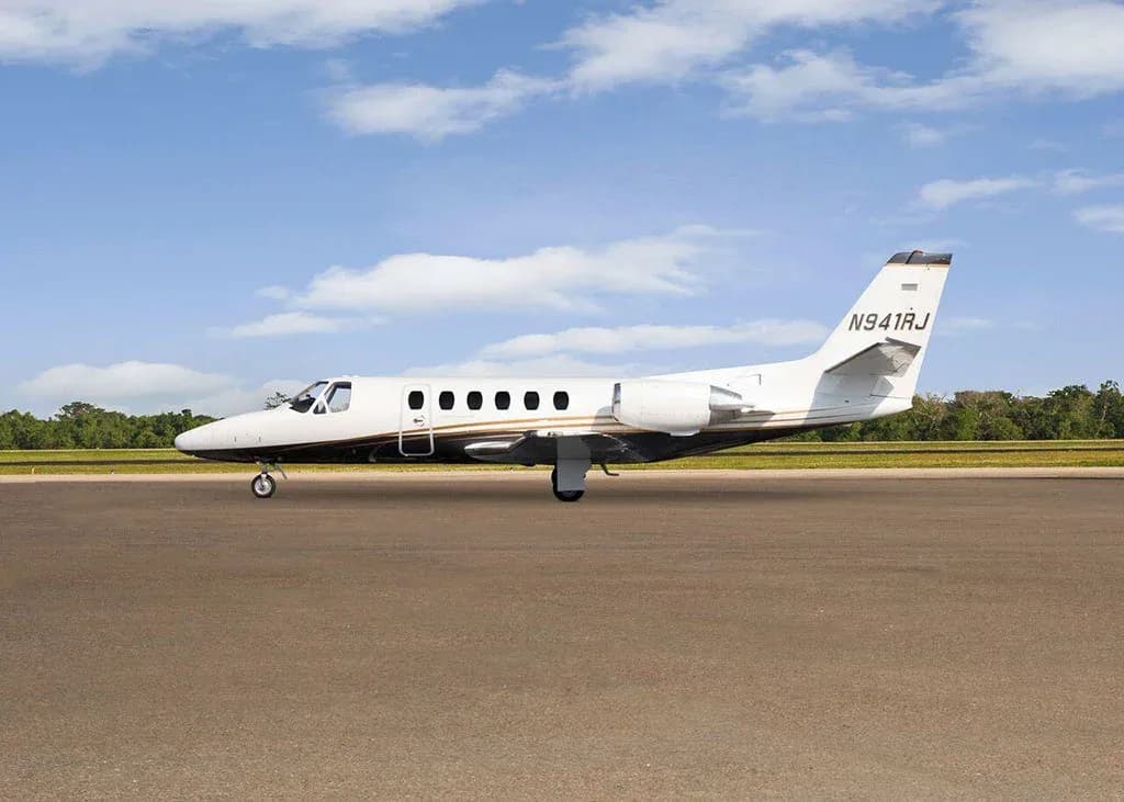A white and black Cessna Citation business jet N941RJ with gold stripes is parked on a tarmac under a partly cloudy sky, viewed from the side.
