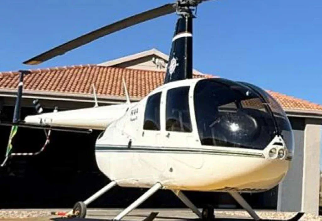 A close-up front view of a white Robinson R44 Raven II helicopter parked in front of a building with a red tiled roof.