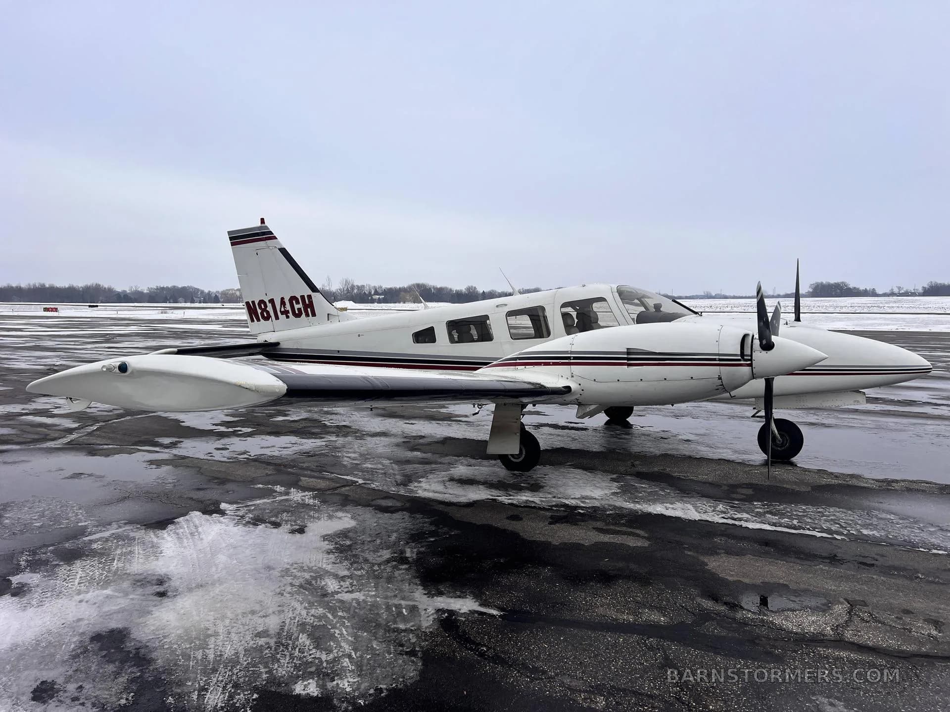 A side view of a white twin-engine Piper aircraft with maroon and black stripes, tail number N814CH, parked on a wet tarmac with snow.