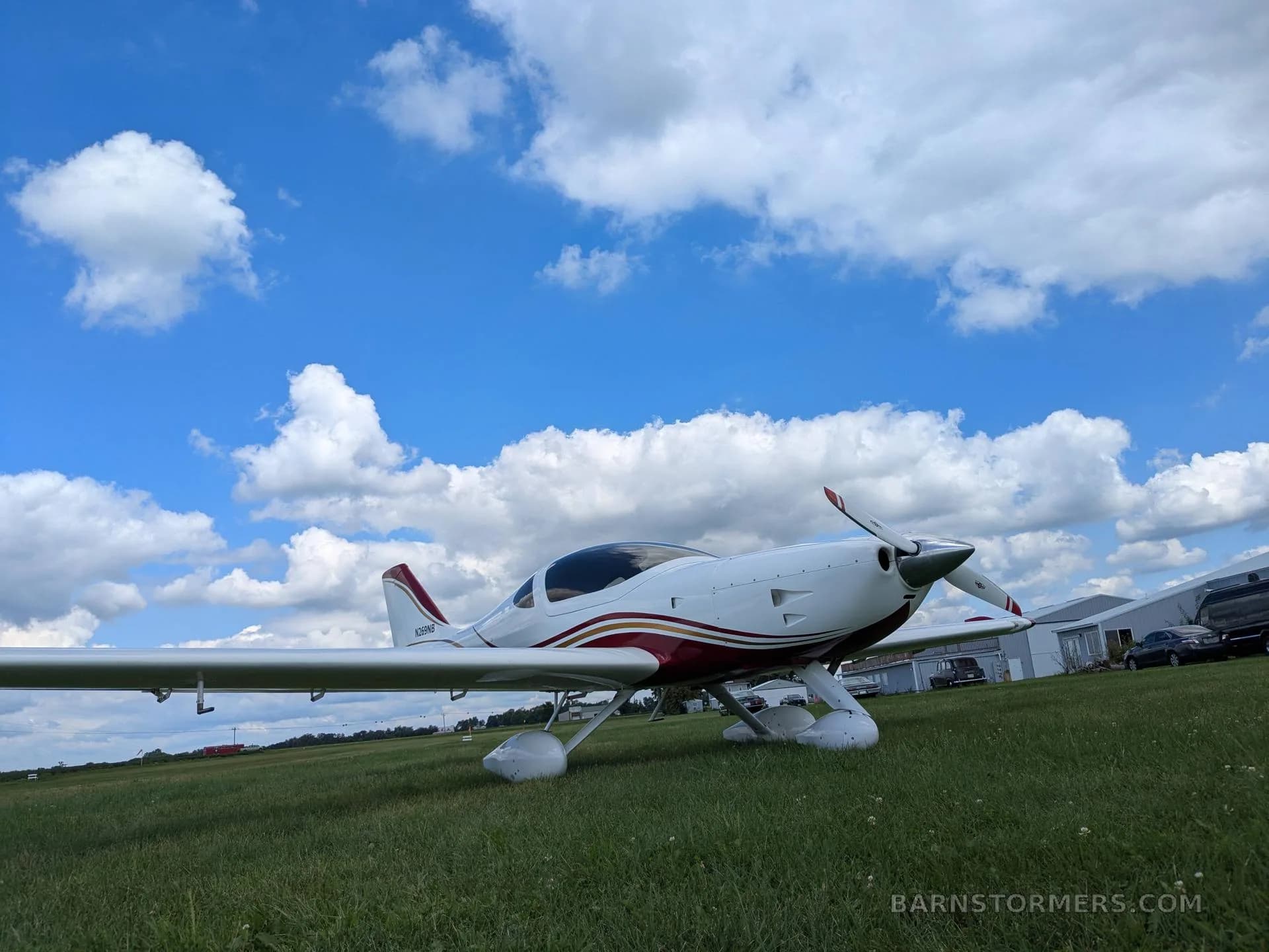 A white and red aircraft with a propeller is parked on a grassy field with buildings in the background.