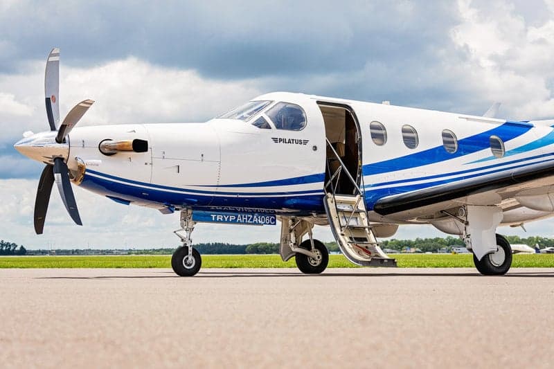 A Pilatus PC-12 aircraft with a blue and white paint scheme is parked on the tarmac with the door open.