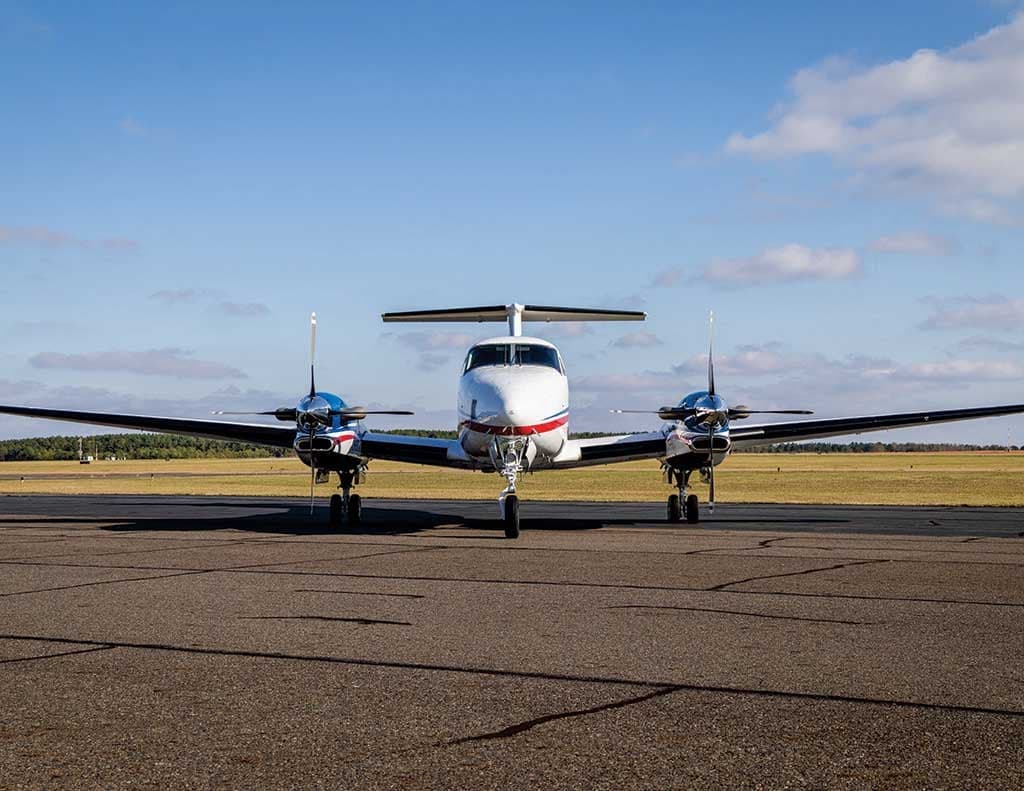 Front view of a white Beechcraft King Air 360 with blue and red stripes on a tarmac under a blue sky.