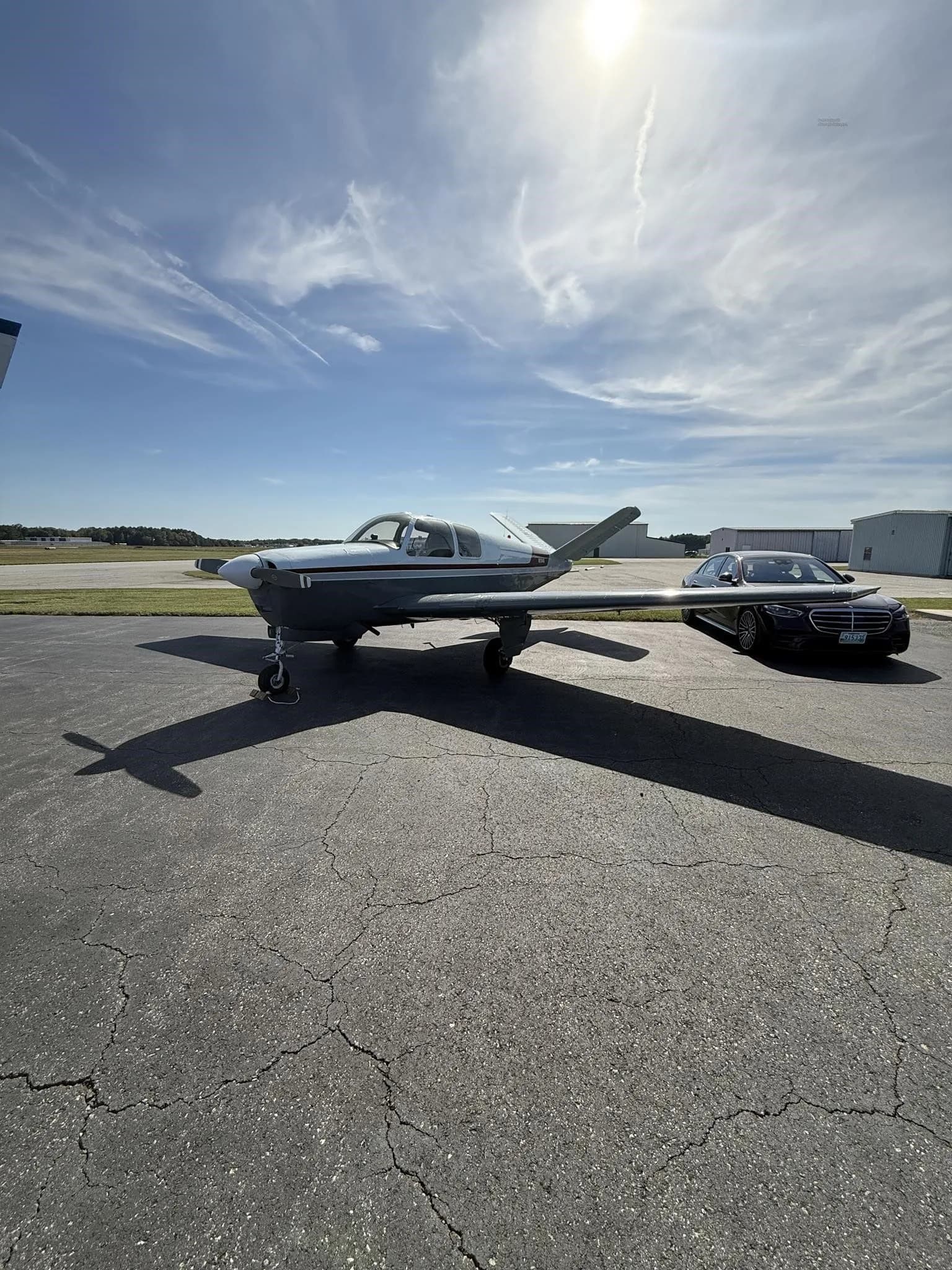 The N2194D Beechcraft Bonanza V-tail aircraft, with a white and grey paint scheme and red stripe, parked on an airfield tarmac next to a black car.