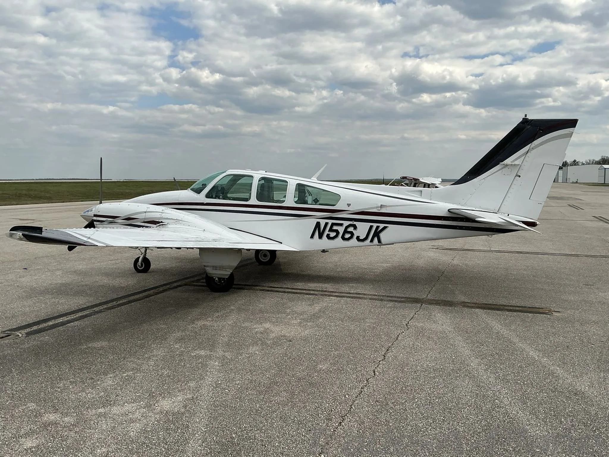 A side view of a white twin-engine propeller aircraft with N56JK tail number, black and maroon stripes, parked on an asphalt tarmac.