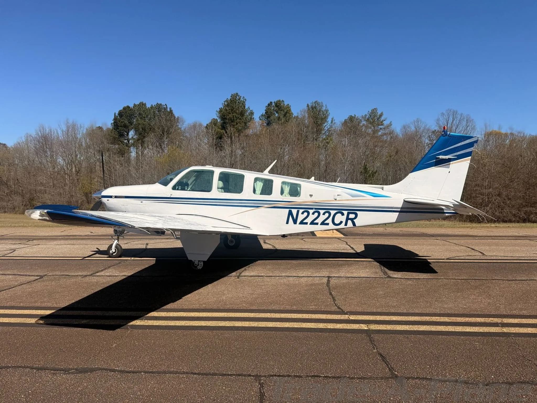 A white Beechcraft Bonanza N222CR with blue and gold stripes parked on an asphalt runway under a clear blue sky.