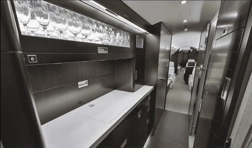 The galley area of a private jet, featuring dark wood cabinetry, a counter, and a display of glassware.